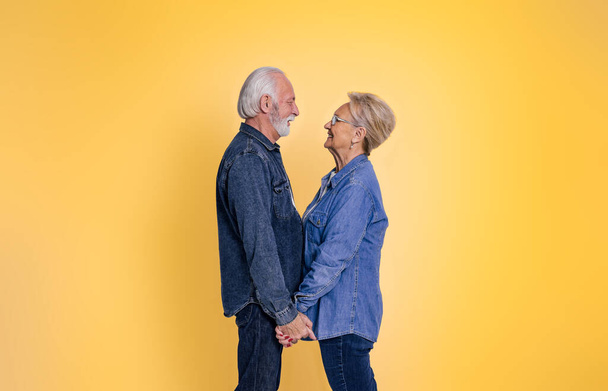Boczny widok kochającego starszego mężczyzny i kobiety trzymających się za ręce i romantycznie patrzących na siebie stojących na żółtym tle - Zdjęcie, obraz