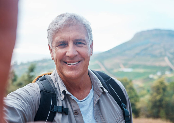 Selfie, hiking and portrait of old man on mountain for outdoor adventure, freedom and wellness in nature. Retirement, traveling and happy senior male take picture on hike, trekking and summer holiday. - Photo, Image