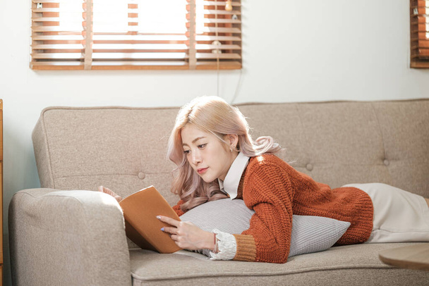 Portret van een Aziatische vrouw die een boek leest in de woonkamer. 's Ochtends warme koffie of chocolademelk drinken. Hoge kwaliteit foto. - Foto, afbeelding