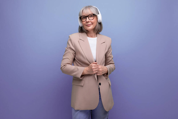middle-aged woman in a stylish jacket listens to music in large headphones on a bright background with copy space. - Photo, Image