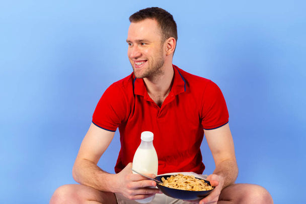 A young, handsome, healthy man in a red polo shirt is holding a bowl of cereal or breakfast and a bottle of milk. Isolated on blue background. - Foto, imagen
