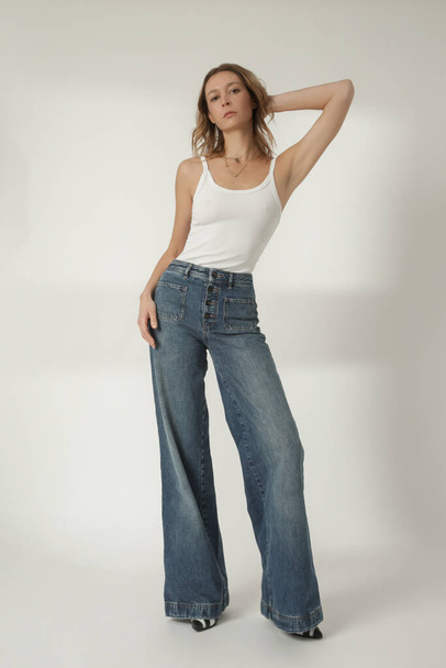 Fashion portrait of young woman in white tank top and wide leg blue jeans on the white background - Fotó, kép