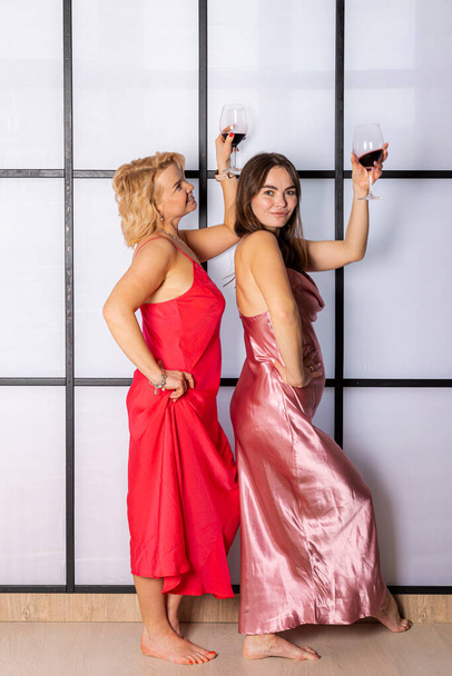 Two charming women drinking wine and having fun, dancing, enjoying party, hand up. Fashionable skinny girls in sexy shiny dresses with paillettes, pretty wavy hairstyle. - Photo, Image