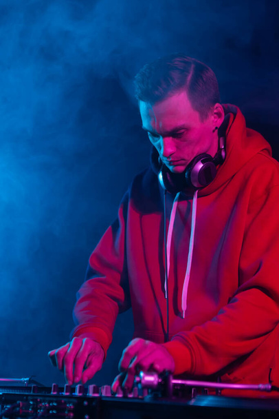 Cool young hip hop DJ in red hoodie mixing vinyl records on a party in night club. Professional disc jockey plays set in smoke on stage - Valokuva, kuva