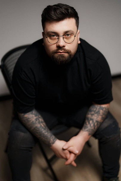 Contenuto giovane modello maschio barbuto plus size in abito nero e occhiali con braccioli tatuati seduti su sedia su sfondo grigio mentre in studio - Foto, immagini