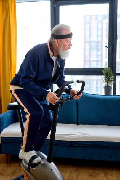 caucasian aged man in mid 70s exercising on stationary bike at home alone. Active mature male in sportswear during morning workout routine. in living room, man lead healthy lifestyle - Photo, image