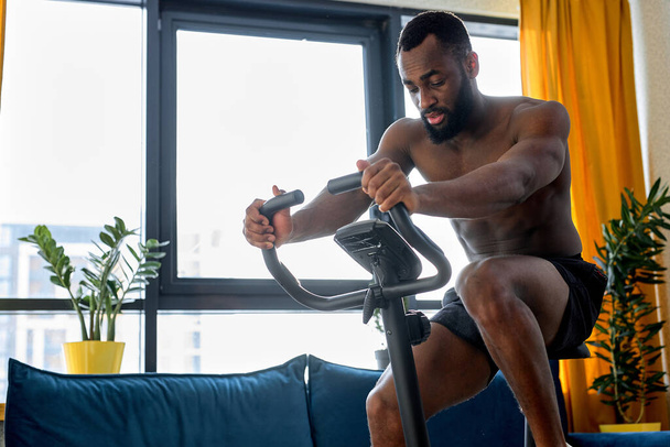confident black man working out on exercise bike or stationary bike at home alone. fitness man with naked torso engaged in cardio training. Sport, workout, healthy lifestyle concept. - Photo, image