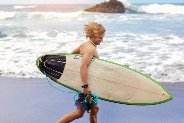 Fit νεαρός surfer άνθρωπος με σγουρά ξανθά μαλλιά με σανίδα του σερφ πηγαίνει από τον ωκεανό διασκεδάζοντας κάνοντας ακραία θαλάσσια σπορ, σέρφινγκ. Ταξίδι και υγιεινό τρόπο ζωής έννοια. Αθλητικός προορισμός ταξιδιού - Φωτογραφία, εικόνα