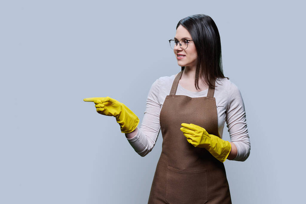 Young woman in rubber glove apron pointing presenting with her hand at empty copy space for text, on grey background. Positive confident female cleaning service worker, smiling housewife - Photo, Image