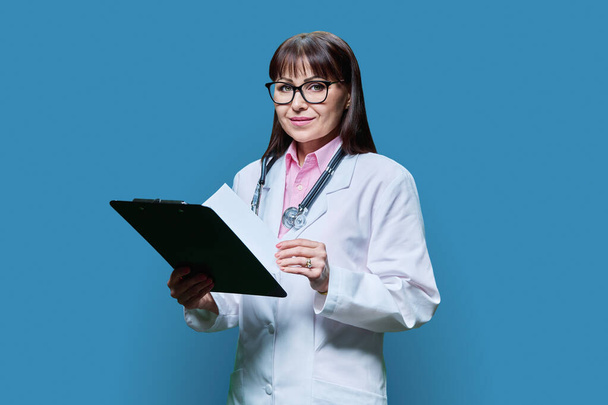 Female doctor with clipboard on blue studio background. Middle aged smiling female medic in white coat with stethoscope looking at camera. Medicine, healthcare, health care, treatment concept - Fotó, kép