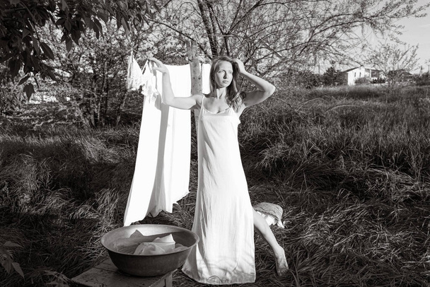 όμορφη κόκκινη κοπέλα με νυχτικό να κρεμάει άπλυτα έξω. χωριό γυναίκα που εργάζονται στην ύπαιθρο. Χαριτωμένο κορίτσι με φόρεμα πλύσιμο λευκά ρούχα σε μεταλλική λεκάνη στην πίσω αυλή, κρέμεται πλυντήριο σε σχοινί ρούχων - Φωτογραφία, εικόνα