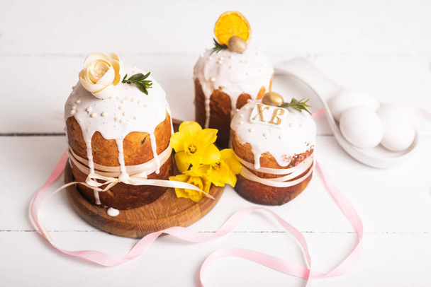 黄色の水仙と白い木製の背景に繊細で軽やかでモダンなデザインの卵を持つイースターケーキ。イースターのコンセプト。イースターの伝統的な食べ物。イースターシンボル - 写真・画像