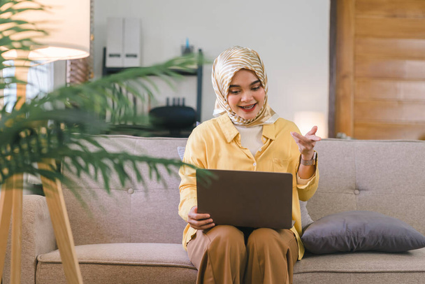 A la mujer musulmana se le muestra una videollamada en su computadora portátil en una teleconferencia con sus compañeros de trabajo. Ella está sentada en un sofá en su sala de estar y se puede ver sonriendo y haciendo gestos hacia la cámara - Foto, imagen