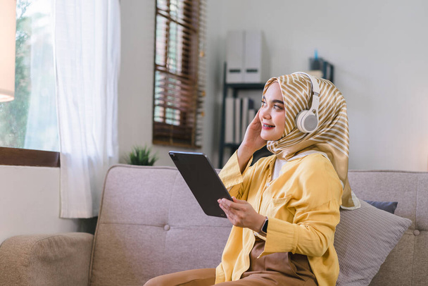 自宅のタブレットを使用してヒジャーブを身に着けているアジアのイスラム教徒の少女の肖像画、リビングルームのソファに座っている間、ソーシャルネットワークをメッセージングまたはブラウジング - 写真・画像