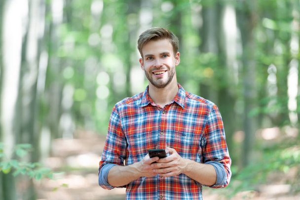 glad man messaging on phone outside. man messaging on phone wearing checkered shirt. photo of man messaging on phone in the forest. man messaging on phone outdoor. - Foto, Imagem