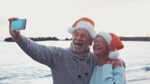 videomateriaalia kahdesta onnellisesta ja aktiivisesta vanhuksesta tai eläkeläisestä joulupukin hatuissa juhlimassa joulua rannalla auringonlaskun aikana - Materiaali, video