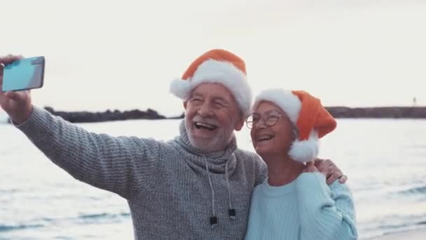Съемка двух счастливых и активных пожилых людей или пенсионеров в Санта-Шапке, празднующих Рождество на пляже во время заката - Кадры, видео