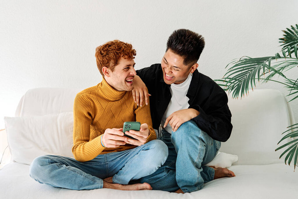 Porträt eines glücklichen homosexuellen Paares, das mit einem Handy auf dem Sofa sitzt - Foto, Bild