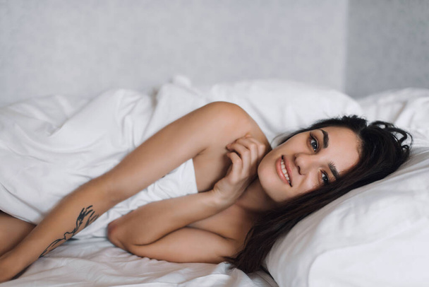 sexy feliz desnudo hermosa chica morena sonriendo en la cama bajo una manta en el dormitorio - Foto, imagen