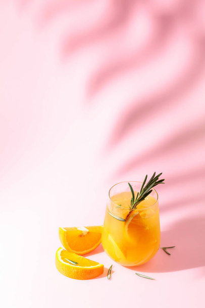 Summer cocktail with orange, rosemary, and ice. Drink on pink background with palm leaf shadow. Summer, tropical, fresh cocktail concept. - Foto, imagen