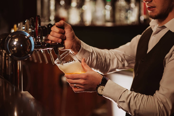 hands of bartender pour into glass draught light foamy beer from tap in bar - Фото, зображення