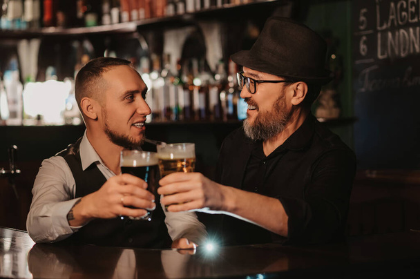 two happy smiling bartenders clink glasses of draft beer at bar counter - Фото, изображение