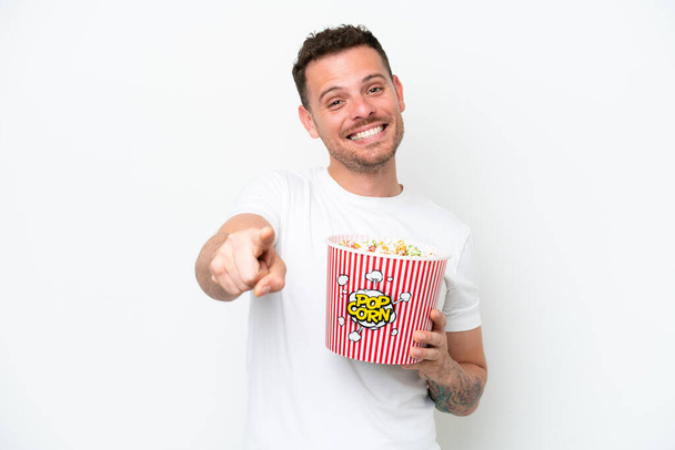 Young caucasian handsome man isolated on white background holding a big bucket of popcorns while pointing front - Photo, Image