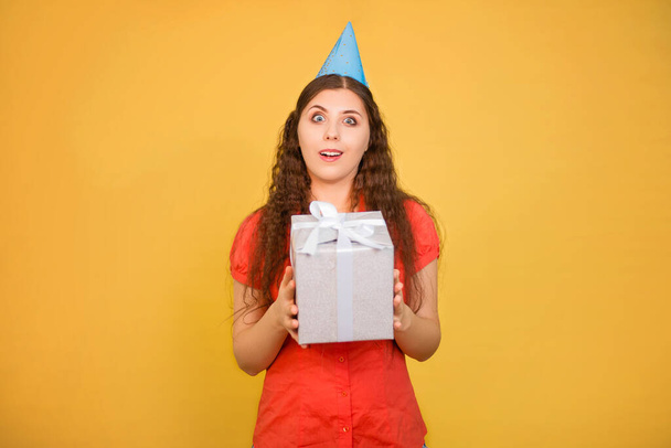 Portrait of a young woman in a paper cap with a gift in her hands isolated on a yellow background, birthday concept. - Foto, Imagem
