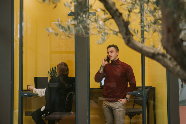 Handsome business man talking on his phone while standing at the little yellow room at the co working space - Photo, Image