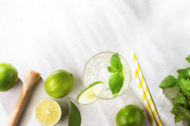 Flat-lay of mojito cocktail with lime, fresh mint and ice on white texture background with shadows. Summer refreshment citrus drink. Top view and copy space. - Foto, Imagem