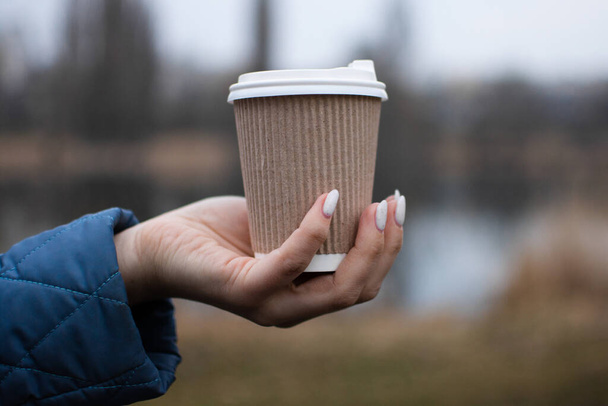 Πίνετε καφέ στη φύση από ένα οικολογικό χάρτινο κύπελλο. Ζεσταίνοντας τα χέρια κρατώντας ένα φλιτζάνι με ένα ζεστό ποτό, περπατώντας ως ένας τρόπος για να ηρεμήσει. - Φωτογραφία, εικόνα