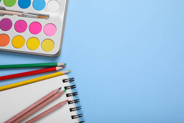 Watercolor palette, colorful pencils and notebook on light blue background, flat lay. Space for text - Fotoğraf, Görsel