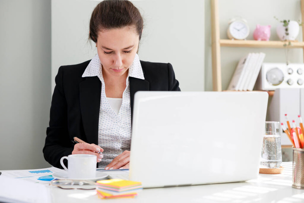 Business woman in classic suit is holding documents, using a laptop while working in home offic - Φωτογραφία, εικόνα
