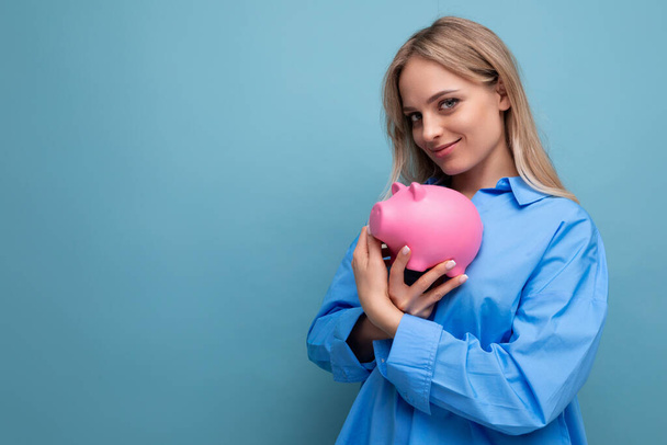 lächelnde blonde junge Erwachsene, die ein Sparschwein umarmt, in dem sie ihre Ersparnisse auf blauem Hintergrund aufbewahrt. - Foto, Bild