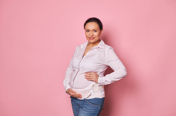 Απομονωμένο πορτραίτο σε ροζ φόντο μιας γοητευτικής Λατινοαμερικάνας εγκύου, φορώντας ορθοπεδικό κοιλιακό επίδεσμο στήριξης, κρατώντας τα χέρια της στην κοιλιά της, χαμογελώντας έντονα, κοιτάζοντας την κάμερα - Φωτογραφία, εικόνα