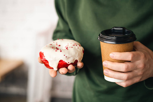Una taza de papel de café y un apetitoso donut de cereza en manos masculinas de cerca, un donut blanco en esmalte con salpicaduras rojas, una deliciosa pausa para el café en el trabajo, desayuno. - Foto, Imagen
