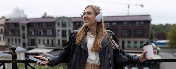 νεαρή χαρούμενη γυναίκα που ακούει μουσική με ακουστικά με ένα φλιτζάνι καφέ στα χέρια της στην πόλη. - Φωτογραφία, εικόνα