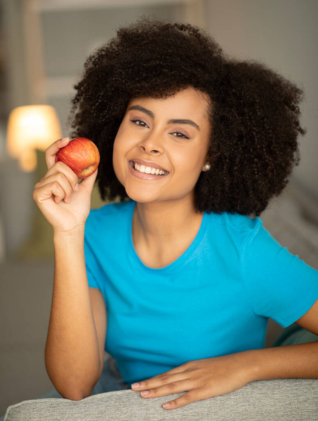 Fröhliche Millennial African American Lockendame genießen Apfel auf dem Sofa im Wohnzimmer Interieur, Nahaufnahme, senkrecht. Gesundheitsfürsorge, richtige Ernährung, zahnärztliche Hilfe und positiver Lebensstil - Foto, Bild