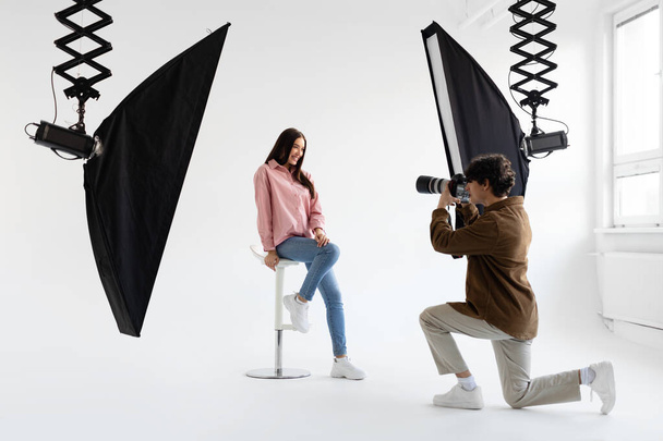 Fotografo professionista scattare foto di giovane donna in abbigliamento casual, avendo photoshoot in studio moderno con attrezzature di illuminazione su sfondo bianco - Foto, immagini