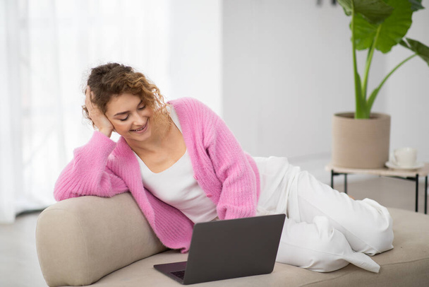 Boldog fiatal nő pihentető laptoppal kanapén otthon, gyönyörű mosolygó nő néz filmeket, vagy böngészés internet a számítógépen, miközben pihen a kanapén kényelmes nappali, élvezi a hétvégi időtöltés - Fotó, kép