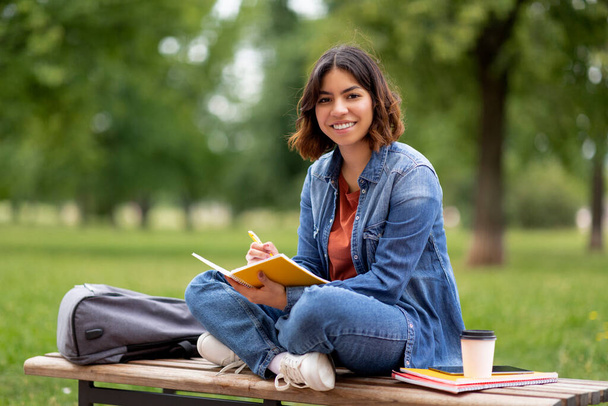 Smiling young middle eastern woman writing in notebook while sitting on bench outdoors, portrait of happy arab female student taking notes to notepad, preparing for exam outside at college campus - Foto, immagini