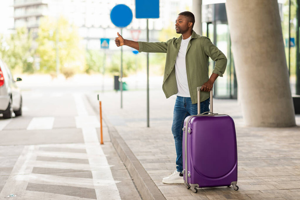 African American Male Tourist Hailing A Taxi Cab Showing Thumbs Up Gesture Standing With Suitcase At Modern Airport Outside. Travel And Transportation Concept. Full Length Shot - 写真・画像