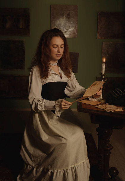 Beautiful woman in vintage clothes reading a letter by candlelight in an art studio - Photo, Image