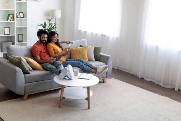 Happy Young Indian Spouses Relaxing With Modern Gadgets On Couch At Home, Married Eastern Couple Resting In Living Room Interior, Man Using Digital Tablet While Woman Browsing App On Smartphone - Фото, зображення