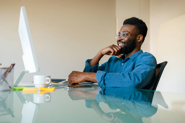 Podekscytowany african amerykański mężczyzna dyrektor generalny siedzi przy biurku przed komputerem, patrząc na monitor i uśmiechnięty, mając wideo rozmowy i przyjemne rozmowy w biurze - Zdjęcie, obraz