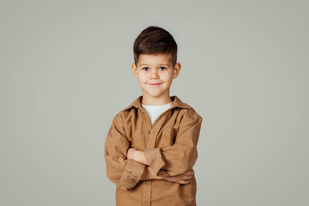Vrolijk zelfverzekerd Kaukasisch 6 jaar oud jongetje in casual met gekruiste handen op borst kijken naar camera geïsoleerd op grijze studio achtergrond. Lifestyle, kindertijd, onderwijs op school, reclame en aanbod - Foto, afbeelding