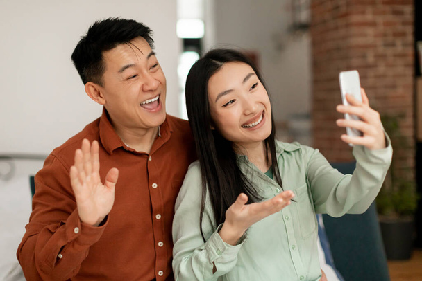 Ενθουσιασμένο κορεάτικο ζευγάρι που χρησιμοποιεί κινητό τηλέφωνο, βιντεοκλήση στο εσωτερικό του σαλονιού, χαμογελώντας στην κάμερα του κινητού. Άνδρας και γυναίκα απολαμβάνουν να μιλούν online με την οικογένεια - Φωτογραφία, εικόνα