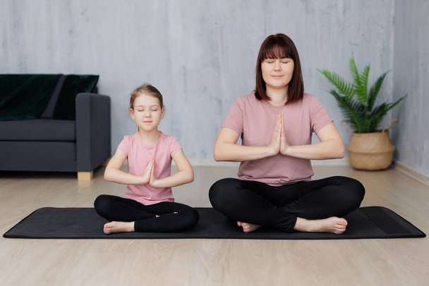 junge Mutter mit kleiner Tochter praktiziert Yoga, sitzt zu Hause in Lotus-Pose auf Yogamatte - Foto, Bild