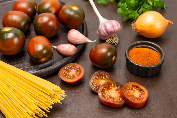 Fresh brown tomatoes and garlic on a plate. Sliced tomato slices, ground red pepper, onion head and pasta on the table. Brown background. - Photo, image