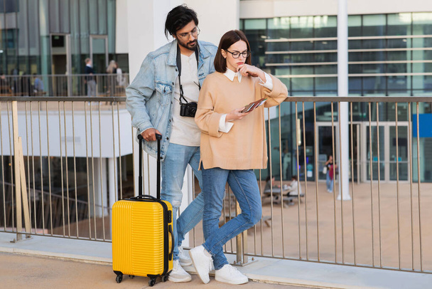 Zoufalý zamyšlený mladý multiraciální pár v neformálních brýlích s pasem, lístky a kufry myslí na letišti ve městě, venku. Cestovní problémy, pozdě na let, dovolená - Fotografie, Obrázek